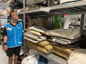 Brenton is smiling while wearing an RSPCA Volunteer uniform. He is standing next to a shelf filled with pet beds. 